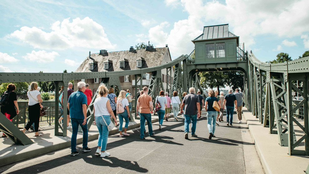 Eine Gruppe Menschen läuft bei einer Führung von Koelnarchitektour zum Hafentag 2023 von Osten nach Westen über die Deutzer Drehbrücke. Die Stimmung ist sommerlich.